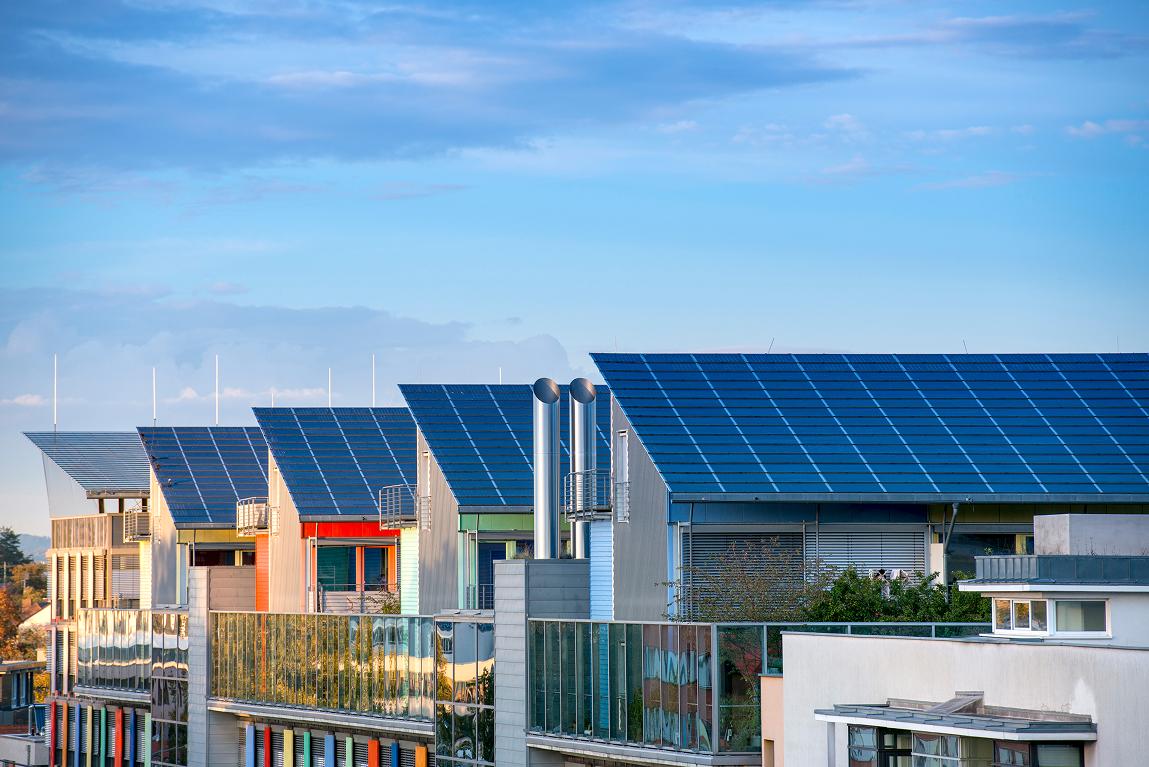 Low-angle view of modern multi-story residential buildings featuring large arrays of blue solar panels installed on their rooftops. The contemporary architecture includes colorful facades, glass balconies, and metallic chimneys, set against a bright blue sky with scattered clouds during what appears to be golden hour.