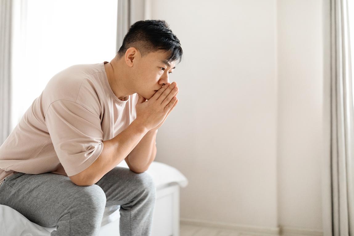A mature Asian man wearing a beige t-shirt and grey pants sits pensively on the edge of a bed in a bright, minimalist bedroom. He leans forward with his hands clasped together near his chin, gazing thoughtfully to the right with a contemplative expression. Soft natural light illuminates the scene from a large window on the left.