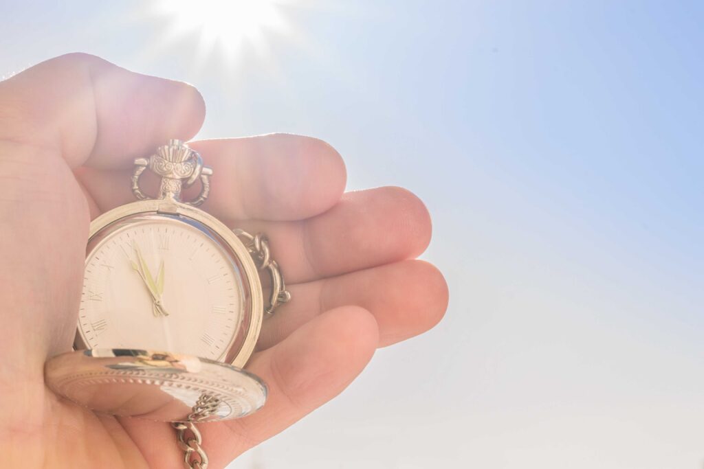 A close-up of a human hand holding an ornate silver pocket watch with its lid open, set against a bright blue sky and a strong sun flare in the background. The watch face shows Roman numerals and the hands pointing near 11 o'clock, with sunlight reflecting off the metallic surface.