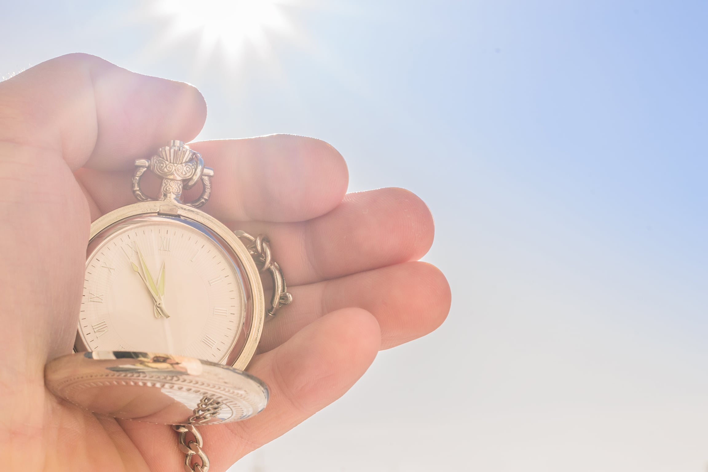 A close-up of a human hand holding an ornate silver pocket watch with its lid open, set against a bright blue sky and a strong sun flare in the background. The watch face shows Roman numerals and the hands pointing near 11 o'clock, with sunlight reflecting off the metallic surface.