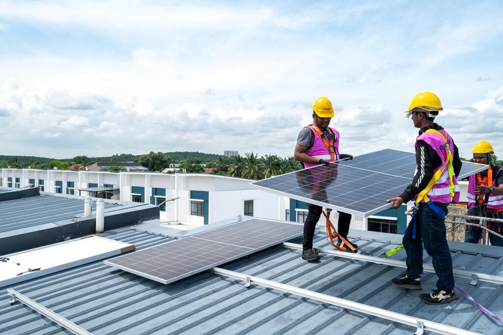 Three solar panel installers wearing yellow hard hats and bright pink safety vests are working on a flat industrial rooftop. Two workers in the foreground carefully carry and position a large black solar panel, while a third worker stands nearby observing. Several solar panels are already laid out on the metal roof. In the background are white and teal buildings, palm trees, and a partly cloudy sky.