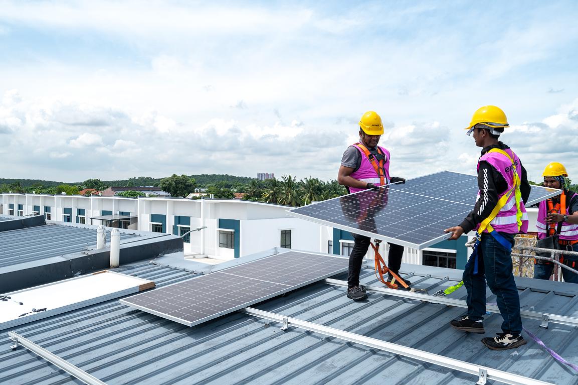 Three solar panel installers wearing yellow hard hats and bright pink safety vests are working on a flat industrial rooftop. Two workers in the foreground carefully carry and position a large black solar panel, while a third worker stands nearby observing. Several solar panels are already laid out on the metal roof. In the background are white and teal buildings, palm trees, and a partly cloudy sky.