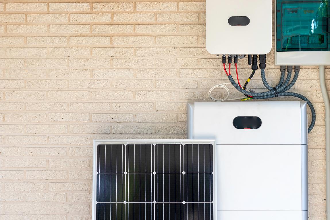 A solar photovoltaic panel leaning against a beige brick wall beside a home battery storage unit, with a wall-mounted solar inverter and electrical wiring above