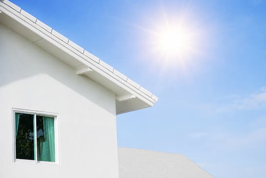 Corner view of a white stucco house with a window and roof eaves against a bright sunny blue sky