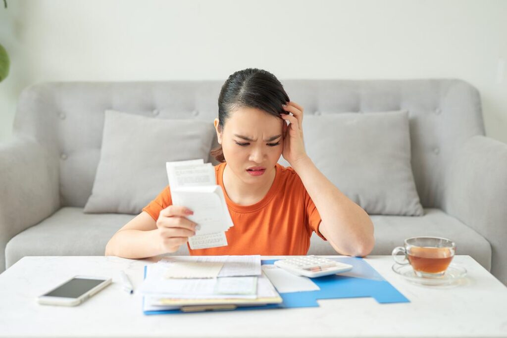 A young woman in an orange shirt sits at a coffee table with a stressed expression, holding a receipt and reviewing utility bills alongside a calculator, phone, and cup of tea