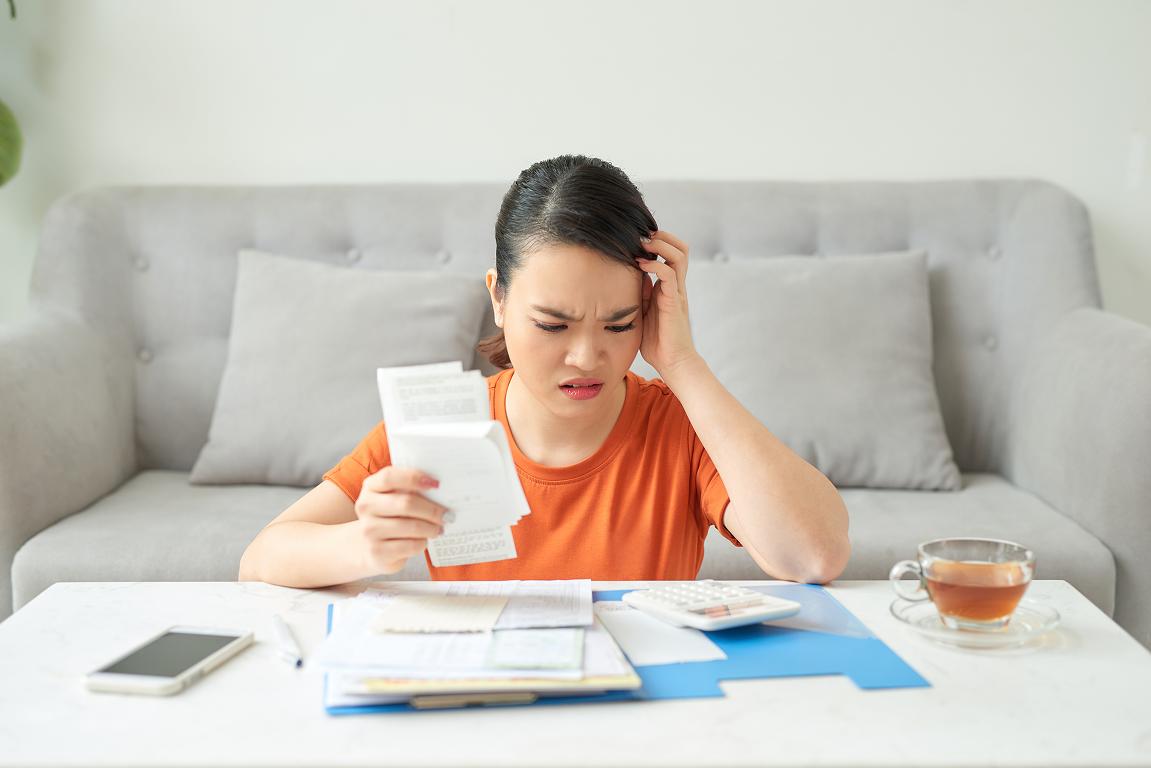 A young woman in an orange shirt sits at a coffee table with a stressed expression, holding a receipt and reviewing utility bills alongside a calculator, phone, and cup of tea